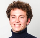 Smiling man with curly hair wearing a dark turtleneck against a light background.