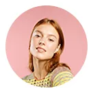 Young woman with shoulder-length light brown hair wearing a yellow patterned top, posing against a pink background.