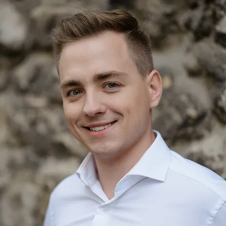 Smiling young man with short light brown hair wearing a white button-up shirt, standing in front of a blurred stone wall.