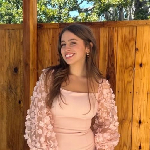 Smiling young woman with long brown hair wearing a light pink dress and floral lace cardigan standing in front of a wooden fence outdoors.